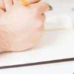 A close up from the side of a mans hands writing in a notebook with a portion of a laptop screen on the right side of the image.