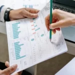 Woman pointing to a paper with a pencil that is held by another person that has graphic representations of statistics.